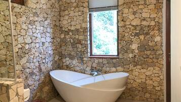 A bathroom with a stone wall, a large bathtub, and a window with blinds.