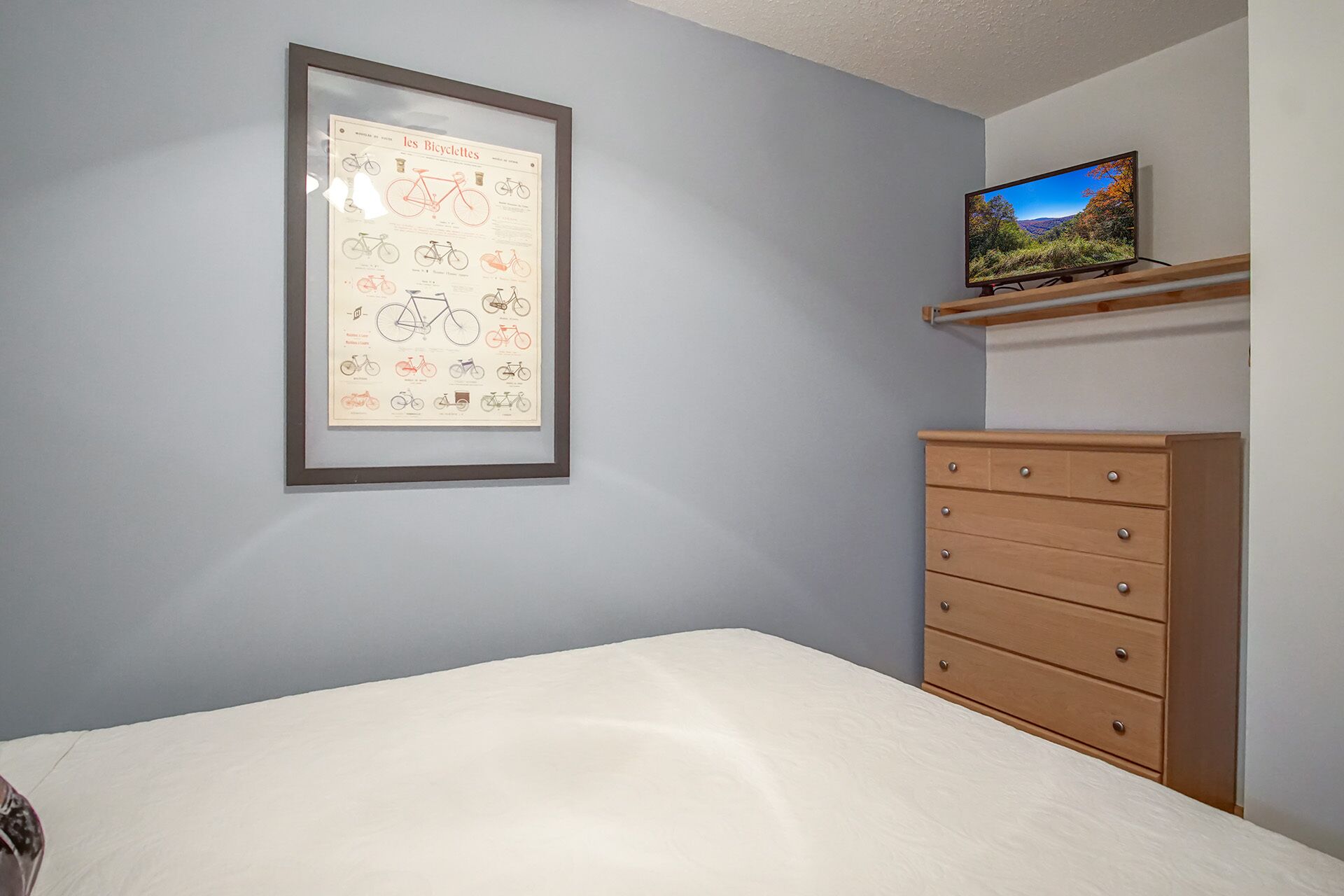 Une chambre à coucher avec un lit, une commode, une affiche encadrée représentant des bicyclettes et un téléviseur posé sur une étagère.