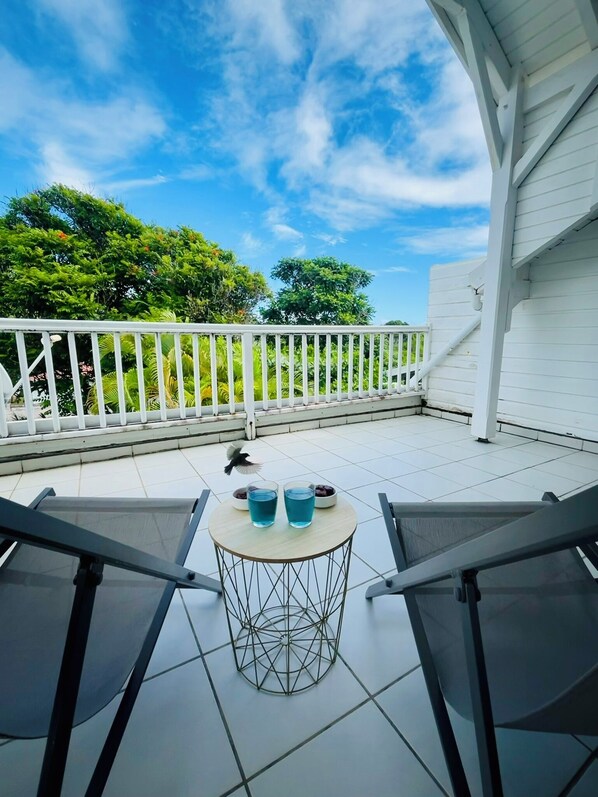 A balcony with a white railing, a round table with two glasses of blue liquid and two bowls of fruit, and a bird flying in the background. - Beautiful, cozy apartment—right on the water (Le Gosier)