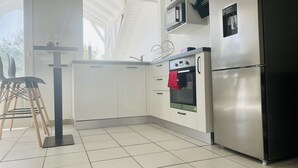 A modern kitchen with white cabinets, a stainless steel refrigerator, a microwave, and an oven. There is a small table with a chair and a window above the sink. - Lovely, cosy apartment - Right on the water (Le Gosier)