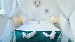 A bed with a green bedspread, white pillows, and a canopy. There are two wall sconces with lights, and two wall mirrors. - Lovely, cosy apartment - Right on the water (Le Gosier)
