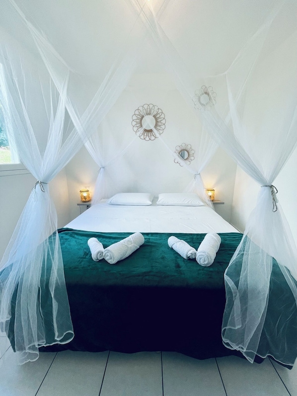 Un lit recouvert d’une couvre-lit verte, avec des oreillers blancs et un baldaquin. Il y a deux appliques murales avec des lampes, et deux miroirs muraux.