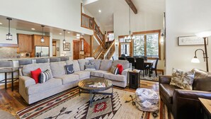 A spacious living room with a large sectional sofa, a coffee table, and a dining area with a wooden table and chairs. A staircase with wooden railings leads to an upper level.