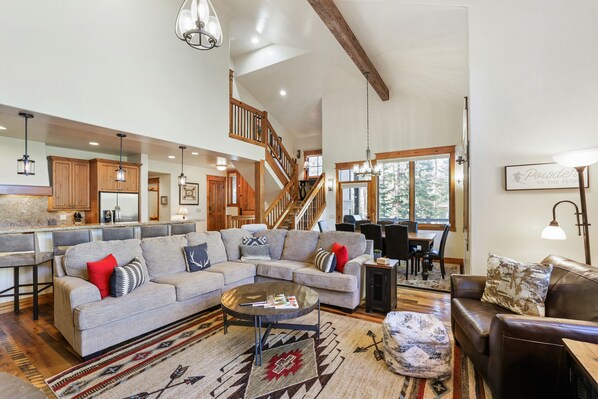 Ein geräumiges Wohnzimmer mit einer großen Eckcouch, einem Couchtisch und einem Essbereich mit Holztisch und Stühlen. Eine Treppe mit Holzwangen führt auf eine höhere Ebene.