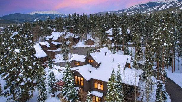 Un albergue cubierto de nieve con varias cabañas, rodeado de un bosque denso y montañas al fondo.
