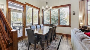 A dining room with a large wooden table and chairs, surrounded by large windows with a view of trees.