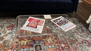 A living room with a dark blue sofa, two pink pillows, and a colorful throw. A glass coffee table holds two magazines. The room has wooden cabinets and a microwave above the stove. - LOCATION!! LOCATION!! LOCATION!! (Oxford)