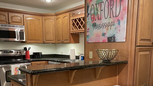 A kitchen with wooden cabinets, a stainless steel microwave, a gas stove, and a bar stool with a white cushion. - LOCATION!! LOCATION!! LOCATION!! (Oxford)