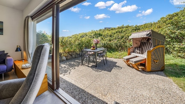 Ein Zimmer mit einer großen Schiebeglasdoor, die auf eine Terrasse im Freien führt. Die Terrasse bietet einen Tisch mit Stühlen, eine Korbsessel-Couch und Blick auf einen Heckenstreifen und den Himmel.