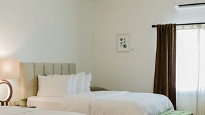 A hotel room with two beds, a green ottoman, a bedside table with a lamp, and a window with curtains. - Wild Iris Room at The Wildflower Inn (Gold Beach)