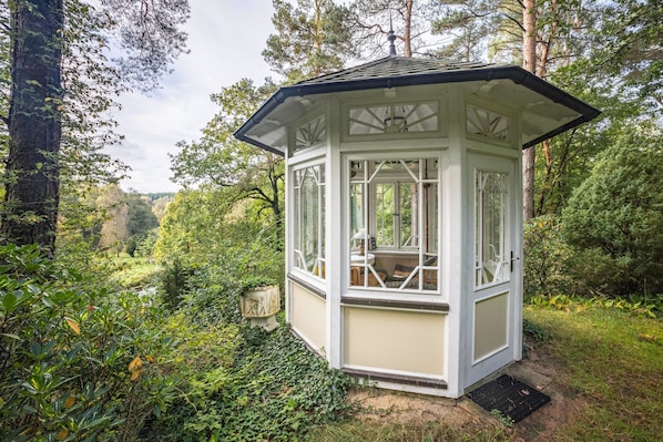 A small, white, glass-paneled gazebo with a peaked roof, surrounded by lush greenery and trees. - 'Romantic Holiday Home' with Garden & Wi-Fi (Gerdau)