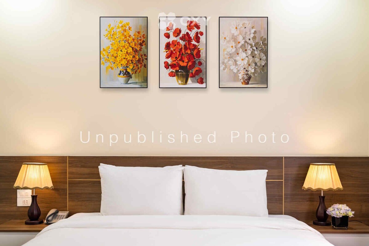 A hotel room with a bed, two bedside tables with lamps, and three framed floral paintings on the wall.