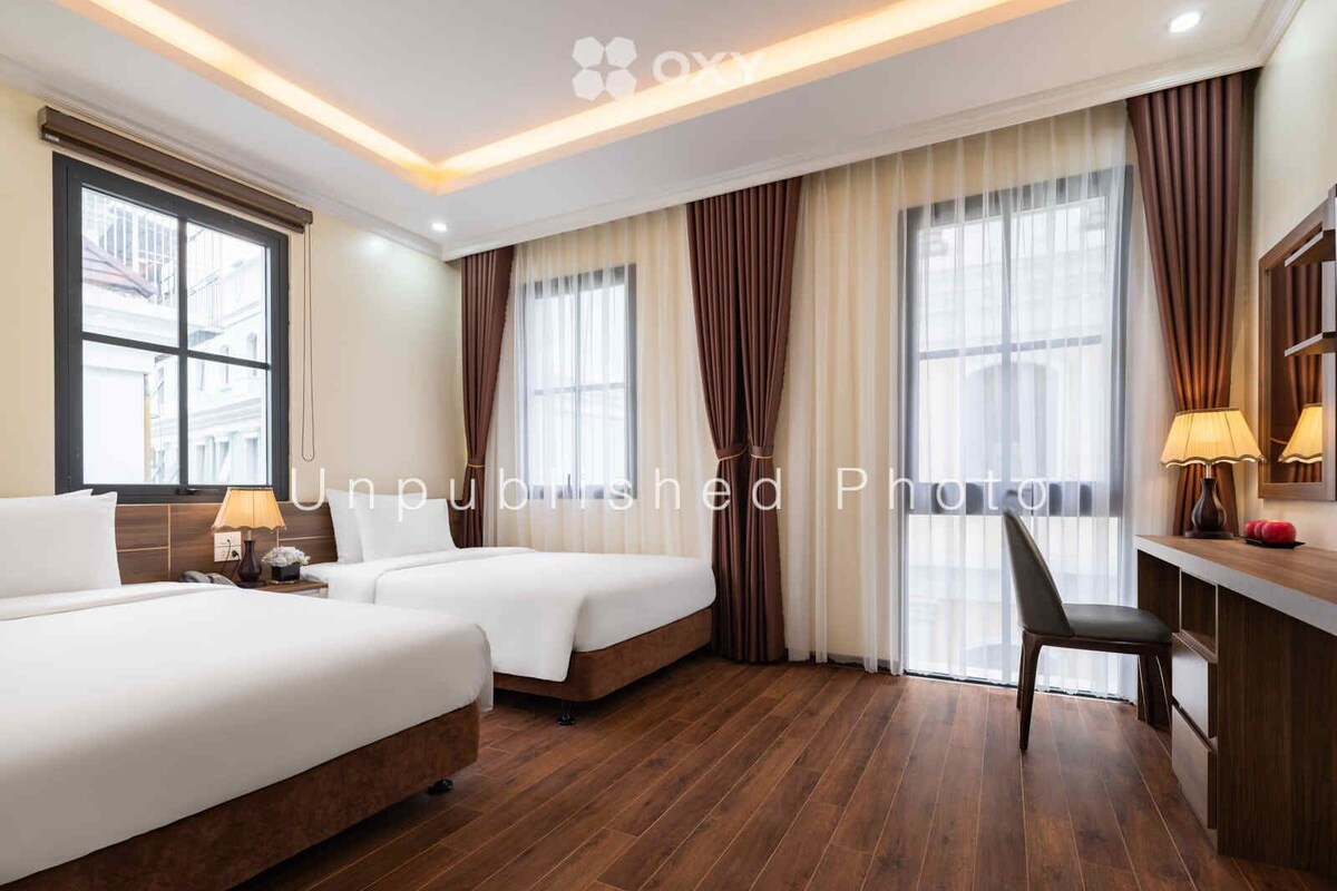 A hotel room with two beds, a desk, and a chair. There are windows with curtains, a lamp on the desk, and a mirror above it.