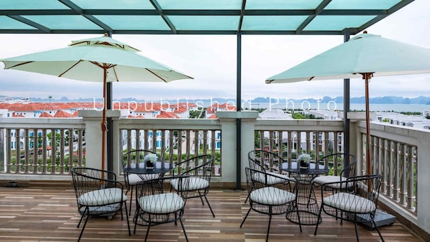 A balcony with two tables and chairs, a glass ceiling, and a view of buildings and water. - Ha Long Essence Classic Hotel (Bai Chay)