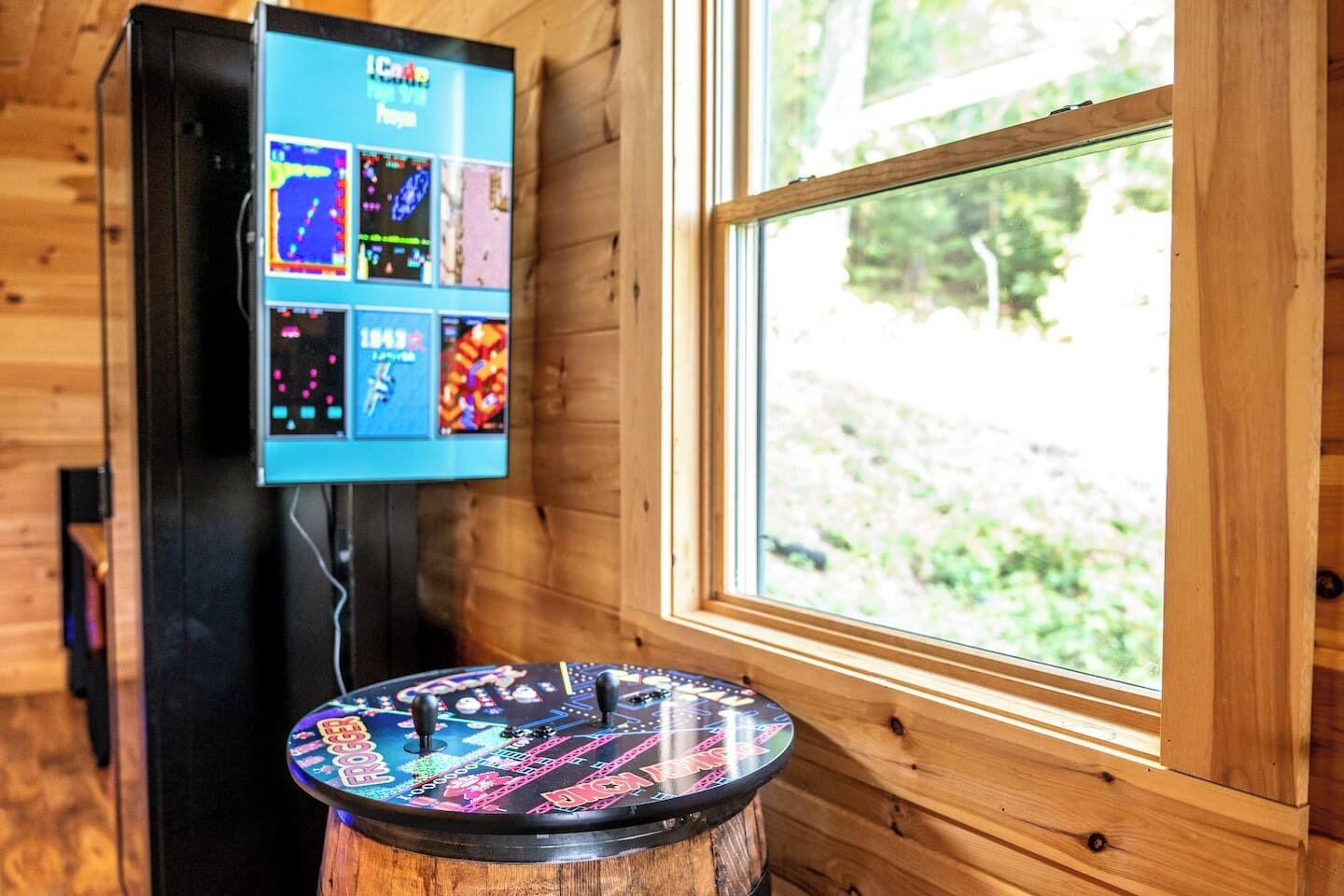 Een houten interieur met een wandgemonteerde scherm dat spelopties toont, een ronde tafel met een kleurrijk tafelblad, en een raam met uitzicht op groen.