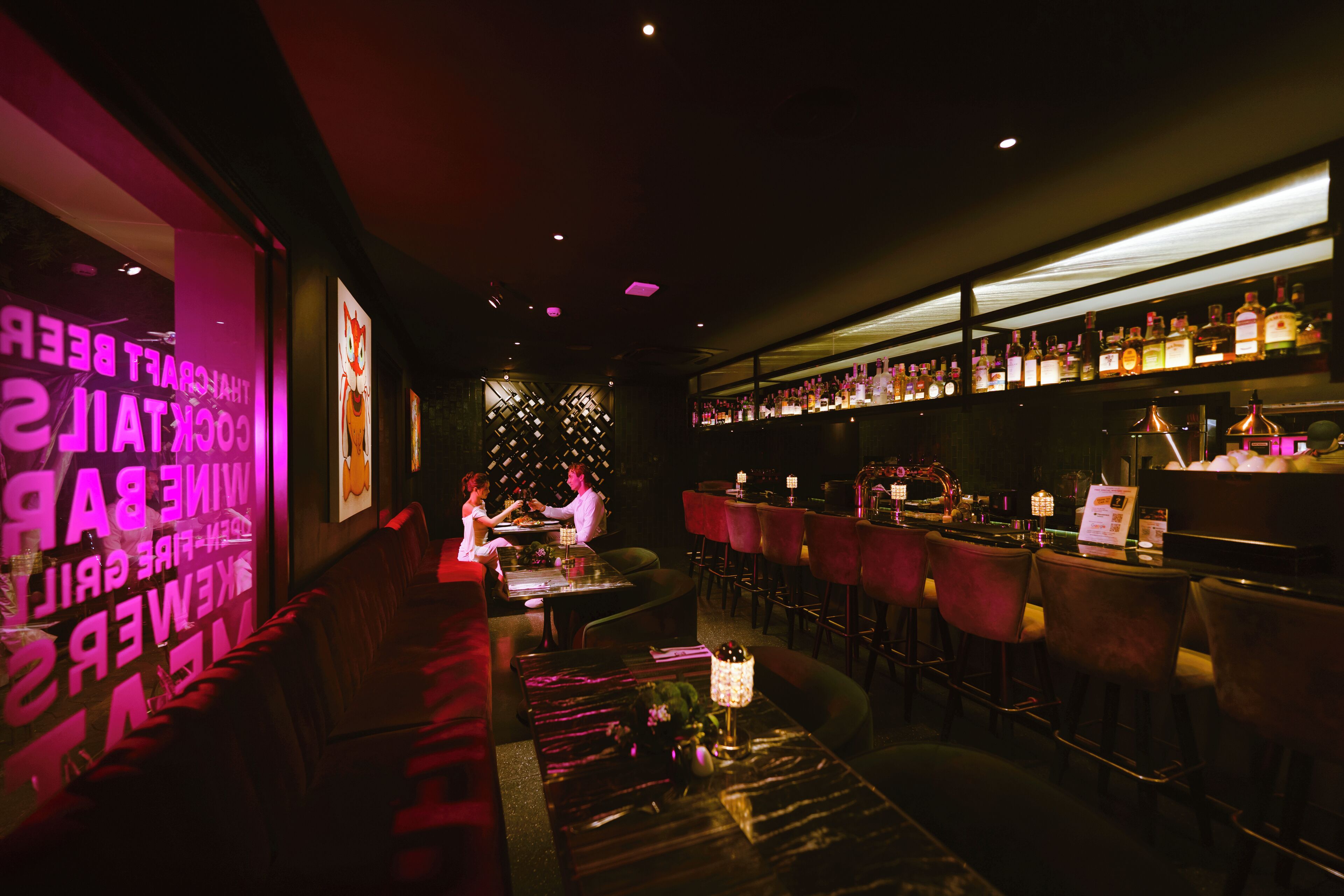 Zona de bar tenuemente iluminada con asientos rojos, barra con botellas y pared decorativa con texto.