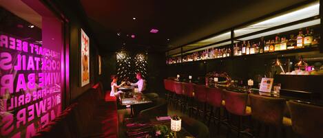 Zona de bar tenuemente iluminada con asientos rojos, barra con botellas y pared decorativa con texto.