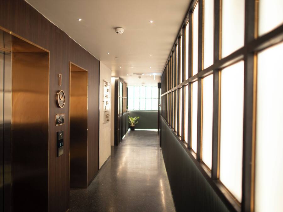 A long, narrow hallway with a modern design, featuring a combination of wooden and metal elements, a potted plant, and a large window on the right side.