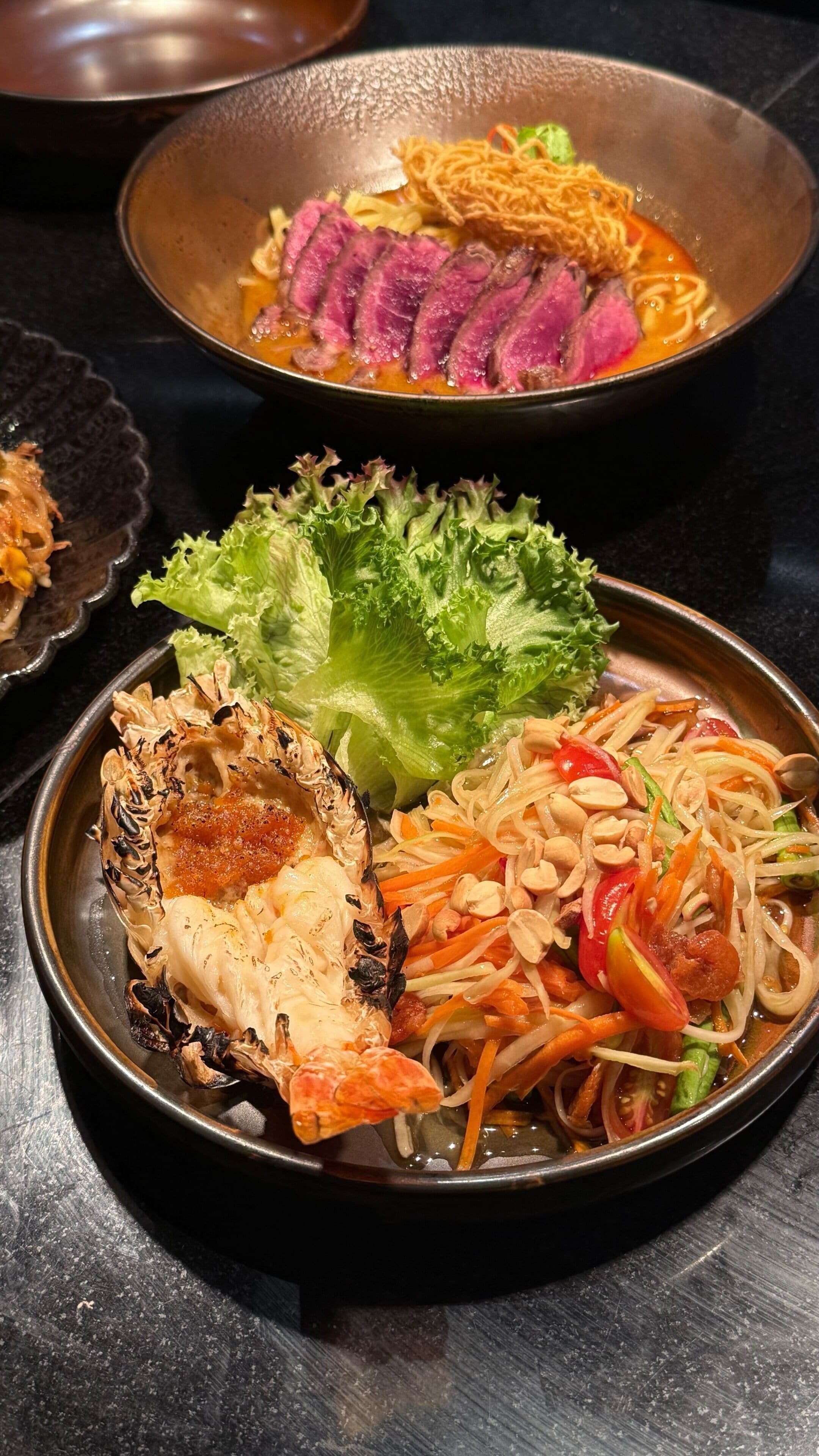 A variety of dishes including grilled lobster, noodles, and a salad with vegetables and peanuts.