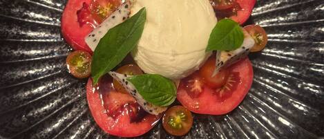 A dish with a central scoop of a creamy white substance, surrounded by slices of red tomatoes and green leaves, on a black plate.