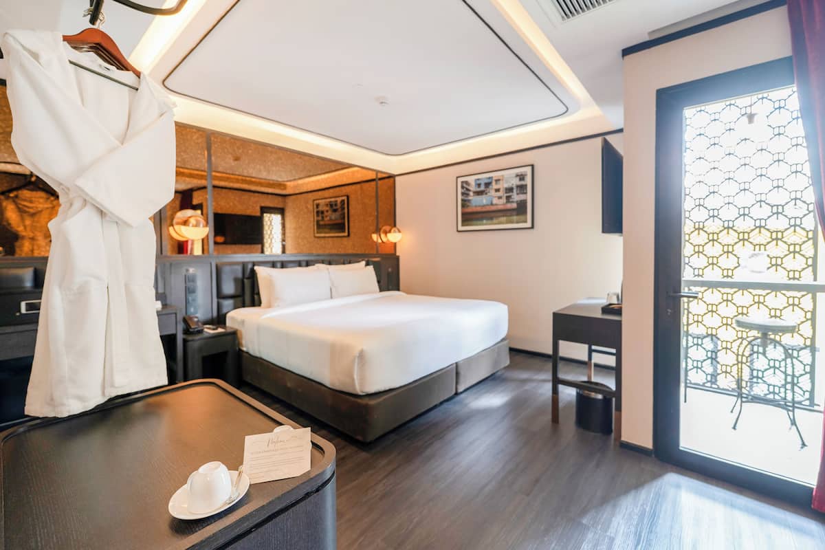 A hotel room with a bed, bedside tables, a desk, a chair, a hanging robe, and a window with a geometric pattern.