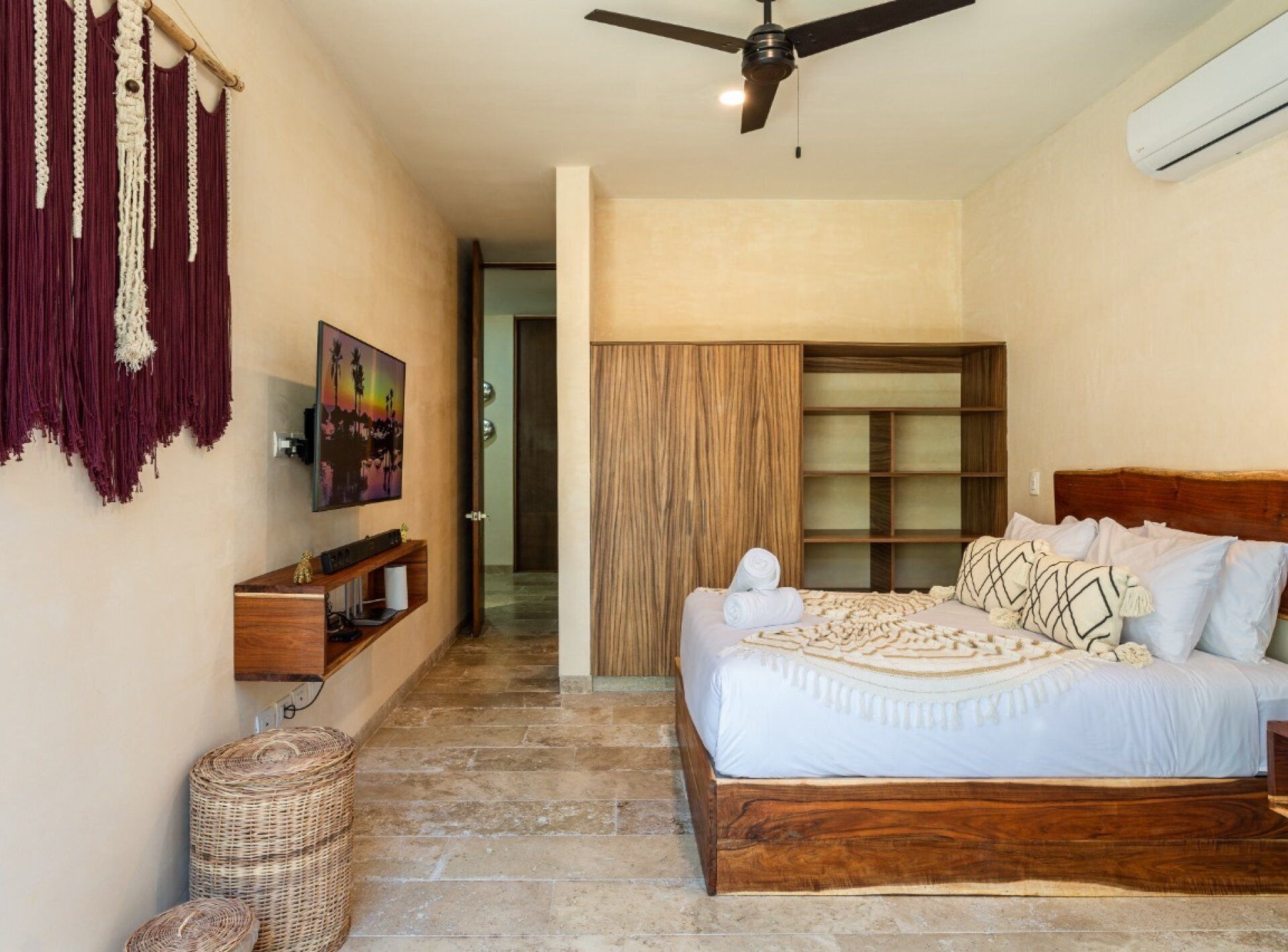 Un dormitorio con una cama de madera, ventilador de techo, televisor y un tapiz de macramé.
