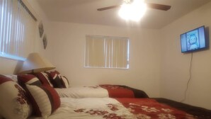 Un dormitorio con cama, ventilador de techo, ventana con persianas y televisor en la pared.