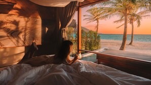 Ein Zimmer mit Bett, Fenster und Blick auf einen Strand mit Palmen.