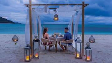 Una pareja cenando en una mesa en la playa, bajo una estructura cubierta con faroles, rodeada de arena y océano.