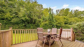 A wooden deck with a table and chairs, surrounded by a wooden fence and lush greenery. - Deluxe lodge with one master double with en suite walk-in shower and one twin. (Cartmel)