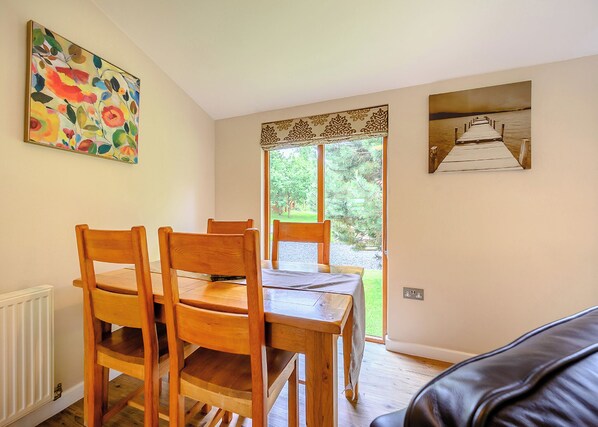 A dining area with wooden chairs and a table, a radiator, a window with a view of greenery, and a painting on the wall.