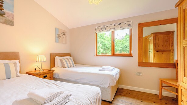A bedroom with two beds, a bedside table with a lamp, a window with a view of greenery, and a wooden wardrobe. - Deluxe lodge with one master double with en suite walk-in shower and one twin. (Cartmel)