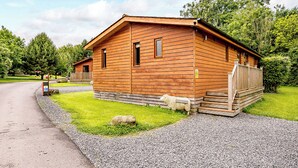 A wooden cabin with a gravel driveway, a wooden deck, and a grassy yard. - Deluxe lodge with one master double with en suite walk-in shower and one twin. (Cartmel)