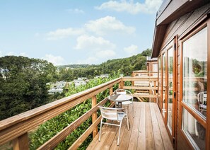 Een houten terras met een tafel en stoelen, met uitzicht op een bebost gebied en gebouwen in de verte.
