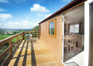 Een houten terras met een tafel en stoelen, een glazen deur naar een kamer met een bank en gordijnen, en een weids uitzicht over groene velden en heuvels.