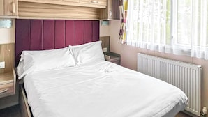 Ein Bett mit weißen Leinen, ein Holz-Kopfteil, ein Nachttisch, ein Fenster mit Vorhängen und eine Heizung.