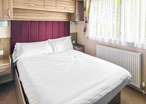 Una cama con sábanas blancas, cabecera de madera, mesita de noche, ventana con cortinas y un radiador.