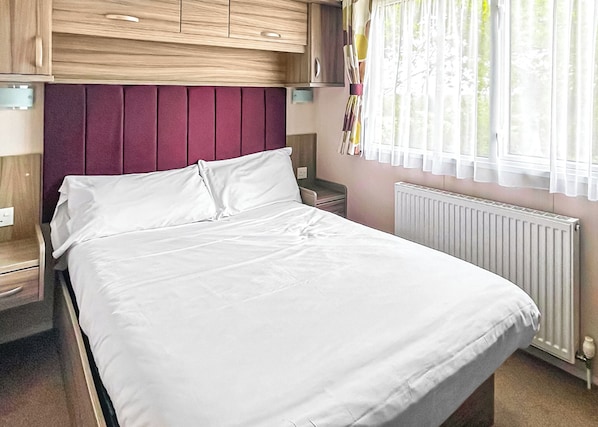 Una cama con sábanas blancas, cabecera de madera, mesita de noche, ventana con cortinas y un radiador.