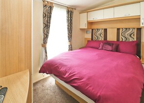 A bedroom with a bed, curtains, a wooden wardrobe, and a small table with a remote control.