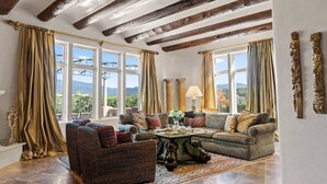 Amplia sala de estar con un sofá grande, un sillón más pequeño y una mesa de centro. La habitación tiene ventanas grandes con cortinas, vigas de madera en el techo y vistas al exterior.