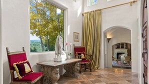 Una habitación con suelo de mármol, un ventanal con vistas a árboles, una chimenea y dos sillones decorativos.