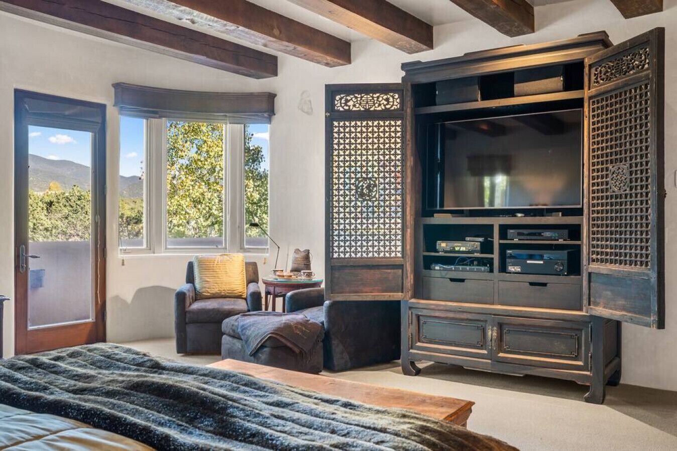 A bedroom with a large bed, a TV stand with a flat-screen TV, a chair, and a window with a view of trees.