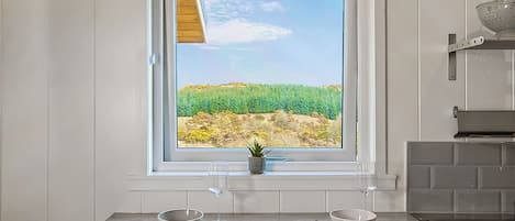 A kitchen with a window view of a green field, a sink, and a colander on the countertop.