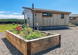 A wooden cabin with a raised garden bed in front, a chimney, and a gravel driveway. - This Studio Lodge is perfect for couples. (Ringford)