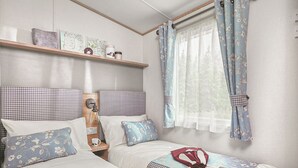 A bedroom with two beds, a shelf with decorative items, a window with curtains, and a bedside table with a lamp. - Thoughtfully designed for comfort and style. (Withernsea)