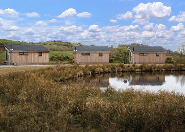 Three wooden cabins with sloped roofs, situated by a calm pond with tall reeds, under a partly cloudy sky.