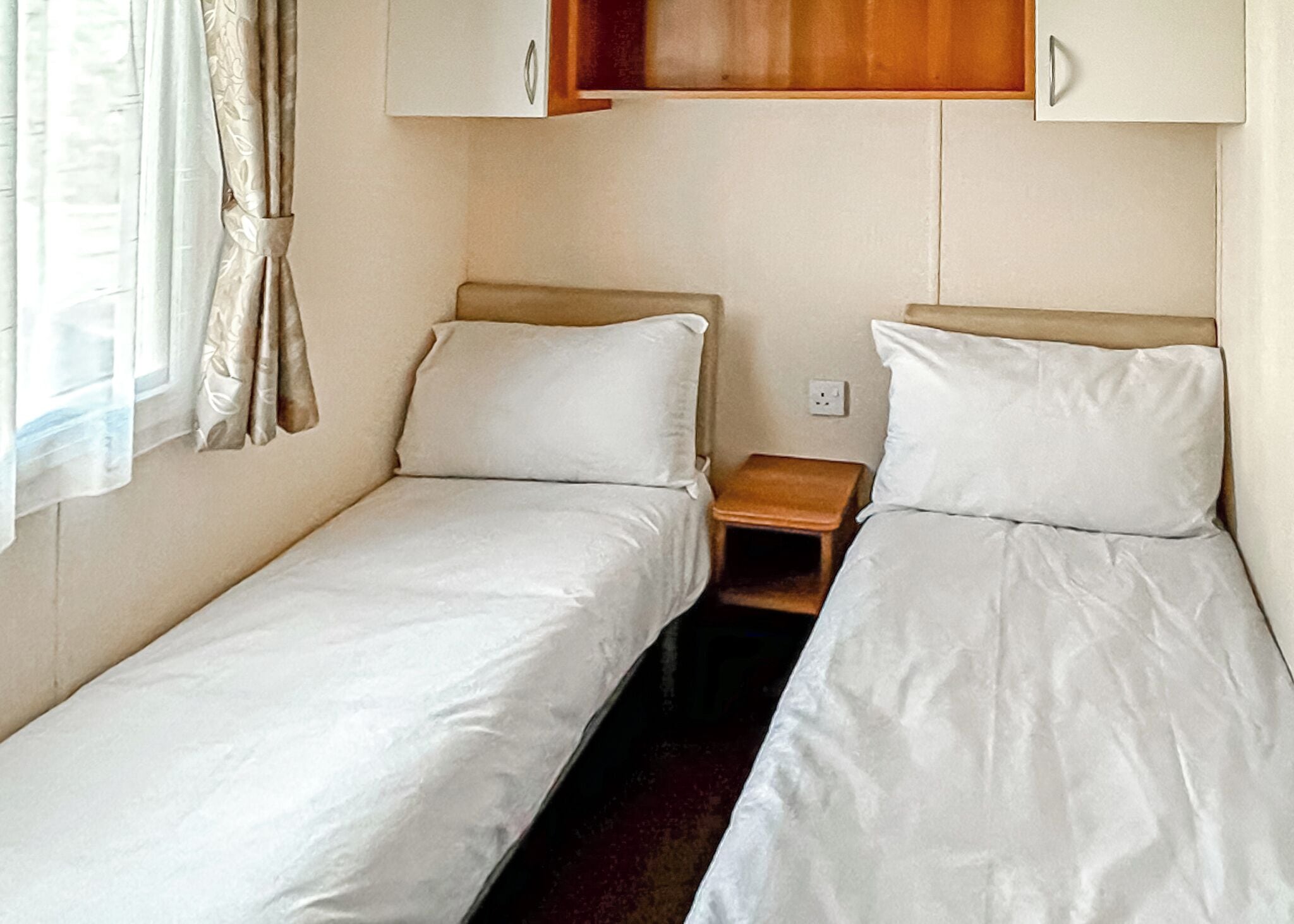 Un alojamiento pequeño de una sola habitación con dos camas individuales, una mesita de noche de madera y una ventana con cortinas.