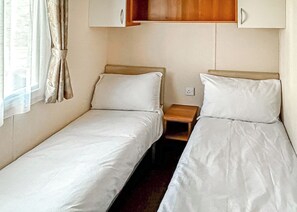 Un alojamiento pequeño de una sola habitación con dos camas individuales, una mesita de noche de madera y una ventana con cortinas.