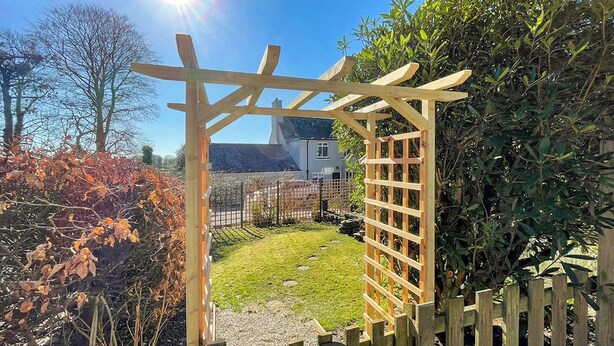 Una estructura de pérgola de madera en un jardín con un sendero de grava y un letrero que indica 'Woodlands Cottage'.
