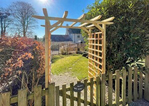 Een houten pergola in een tuin met een grindpad en een bordje met de tekst 'Woodlands Cottage'.